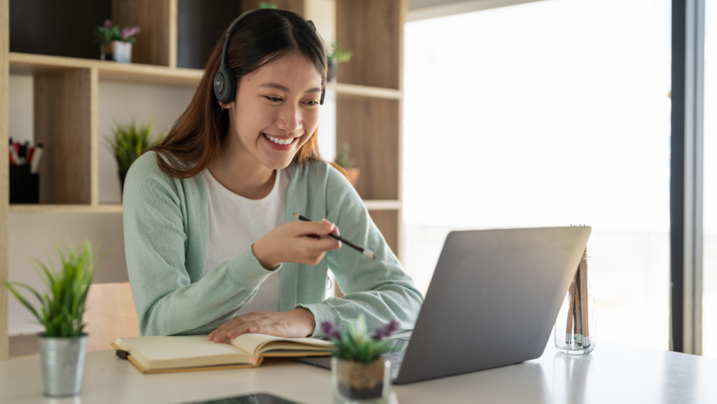 Woman sitting in an office and wearing headphones smiles while pointing a pencil at her laptop